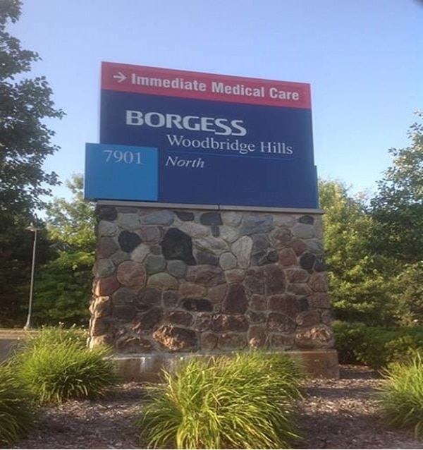 A blue sign on stone. Top of sign, in red with an arrow pointing to the right with text: Immediate Medical Care. Below that, on a blue background with white text: 1st line, BORGESS (large text); 2nd line, Woodbridge Hills (smaller text); and 3rd line, North (smaller text).