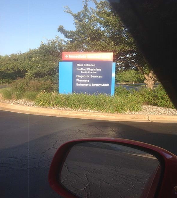 A large blue sign situated in landscaping next to a road or parking lot. Top of the blue sign in red with an arrow pointing to the right with the text: Immediate Medical Care. Below that, on a blue background with white: Main Entrance; ProMed Physicians; Family Practice; Diagnostic Services; Pharmacy; and Endoscopy & Surgery Center.