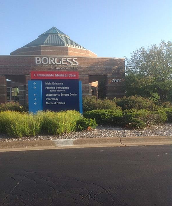 A large blue sign situated in landscaping in front of a brick building. In white text BORGESS with Woodbridge Hills in very small text below. Top of blue sign in red, Immediate Medical Care with left arrow. Below on blue in white text, all the same size, reads: right arrow, Main Entrance; right arrow, ProMed Physicians, Family Practice; left arrow, Endoscopy & Surgery Center; right arrow, Pharmacy; and right arrow, Medical Offices.