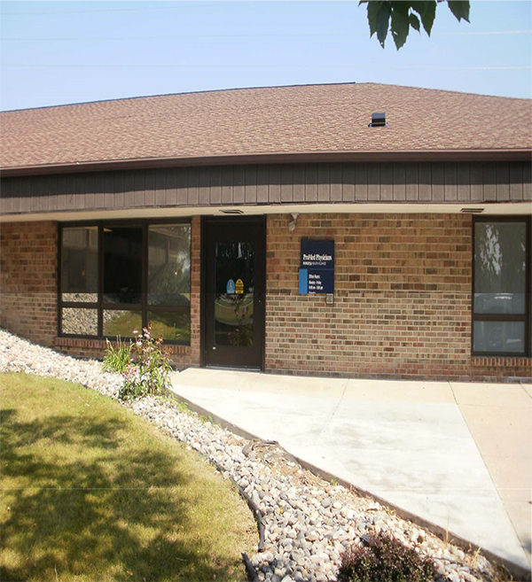 A one-story brick building. Next to the door, a small blue sign with white text: 1st line, ProMed Physicians (large text); 2nd line, BORGESS HEALTH ALLIANCE (smaller text); and 3rd-7th lines, Office Hours: Monday – Friday 8:00 am – 5:00 pm and Saturday 8:00 am – 12:00 pm.